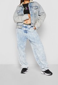 Vaaleansininen farkkutakki, jossa on lampaanvillakaulus, musta lyhyt toppi värikkäällä logolla, ja vaaleat, löysät farkut, jotka on yhdistetty mustiin lenkkareihin.