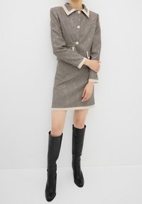 Robe à carreaux marron et crème, avec un col structuré, une fermeture à boutons à l'avant et des manches longues. Associée à des bottes noires jusqu'aux genoux.