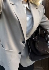 Gai pelēks bleizeris ar izvirzītiem uzvalka apmali, divām melnām pogām un sānu kabatām, kārtots virs baltas un gaiši pelēkas svītrains krekls. Turēta melna rokassoma.