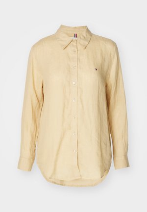 Chemise beige à manches longues avec boutons, col et petit logo brodé sur le côté gauche de la poitrine, sur fond blanc.