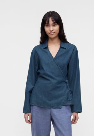 Tânără cu păr drept, lung și închis la culoare, purtând o cămașă de denim albastră în stil tip înfășurare și pantaloni albastre deschis, stând în fața unui fundal simplu.