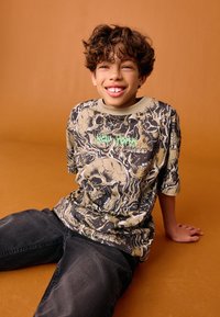 Tricou grafic cu cranii în bej și negru, cu text „New York” de culoare verde deschis. Mâneci scurte și design cu gât rotund.