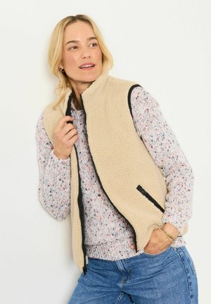 Frau trägt eine beige Fleeceweste über einem gesprenkelten Pullover und blauen Jeans, hält den Kragen der Weste mit einer Hand und schaut zur Seite.