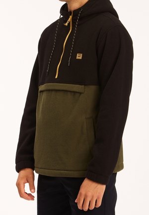 Man wearing black and olive two-tone fleece hoodie with yellow zipper and front pocket, standing against a plain background.
