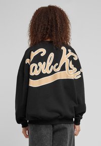 Svart oversized sweatshirt med "Karl K" i stor beige kursiv skrift over ryggen, med en glatt tekstur og ribbede mansjetter.