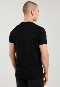 Mees, kelle pea on lõigatud väga lühikeseks, kannab musta lihtsat lühikeste varrukatega T-särki ja tumedaid teksaseid, nähtud tagantpoolt hele taust.