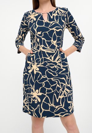 Femme portant une robe bleu marine avec un motif floral abstrait beige et manches trois-quarts, mains dans les poches latérales, sur fond blanc.