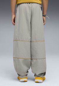 Osoba w luźnych szarych spodniach z żółtym przeszyciem i żółtych sneakersach, stojąca tyłem na szarym tle.