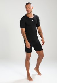 Svart atletisk body med korte ermer og tettsittende design. Har Puma-logo i hvitt på brystet og kort bendekning.