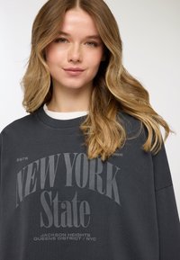 Felpa grigia scuro con vestibilità ampia. Sul davanti è presente un testo audace "NEW YORK State" in grigio chiaro, con testo aggiuntivo sotto. Materiale in cotone.