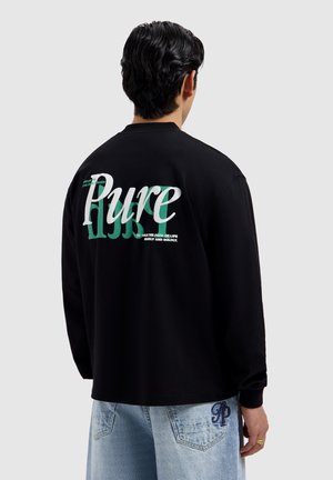 Man in een zwart shirt met lange mouwen met de tekst "Pure" en een groene afbeelding op de achterkant, gecombineerd met lichtblauwe jeans tegen een effen achtergrond.