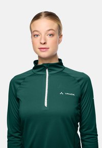 Junge Frau mit zurückgebundenem blonden Haar, die einen grünen Vaude-Sweater mit Viertelreißverschluss trägt, blickt auf einem einfarbigen hellen Hintergrund nach vorne.