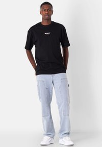 Joven de pie con las manos en los bolsillos, vistiendo una camiseta negra de marca, jeans azul claro y zapatillas blancas sobre un fondo blanco.