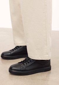 Tenisi negri din piele cu finisaj neted, vârf rotund și talpă plată din cauciuc. Asortați cu pantaloni bej, afișând un design minimalist și șireturi.