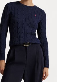 Mörkblå kabelstickad tröja med rund halsringning, prydd med en liten röd logotyp på bröstet, ihop med höga mörka byxor och ett svart bälte.