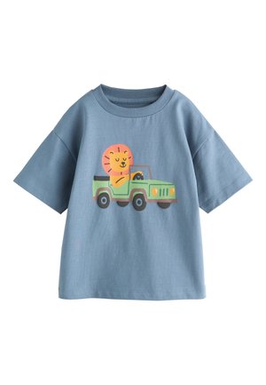 Ung dreng iført blå t-shirt med en løve, der kører en grøn jeep, og beige cargobukser, står mod en ensfarvet baggrund.