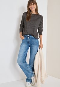 Maglia grigia a maniche lunghe, vestibilità rilassata, abbinata a jeans di denim azzurro a gamba larga. Il modello tiene in mano un giubbotto di pelliccia color crema. Sneakers bianche.