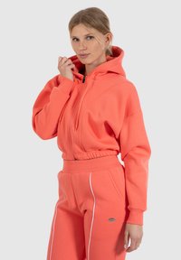 Felpa corallo con zip frontale, coulisse regolabile e vita elastica. Materiale in felpa con piping bianco a contrasto sui pantaloni.