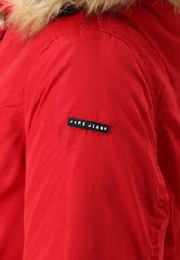 Rød nylonjakke med en kunstpelskrave og et sort mærke med teksten "PEPE JEANS" på venstre skulder. Glat tekstur, puffet design.