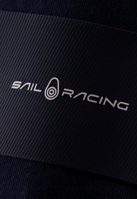 Czarny materiał tekstylny z żebrowaną powierzchnią, na którym znajduje się wyraźne logo "Sail Racing" w białym kolorze, umieszczone centralnie na kontrastowym pasku.