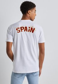 Camiseta de algodón blanca, vista trasera, con el texto en rojo negrita "ESPAÑA" con un efecto desgastado, combinada con jeans de color azul claro.