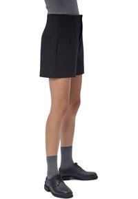 Persona con pantalones cortos negros a medida, calcetines grises, zapatos negros con cordones y una parte superior gris acanalada recortada a la altura de la cintura, de pie sobre fondo blanco.