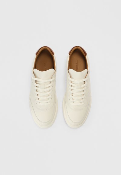 Balti ādas sporta apavi ar gludu tekstūru, apaļu purngalu un brūniem ādas akcentiem papēdī. Aprīkoti ar plakanu auklu aizdari.