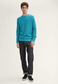 Vaaleansininen neulepaita pyöreällä kaula-aukolla, yhdistetty tummiin farkkuihin. Malli käyttää laivastonsinisiä lenkkareita. Sileä pinta, rento istuvuus.