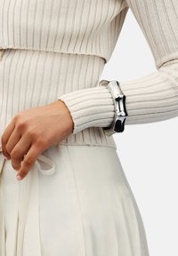 Hand med naturlig hudton som bär ett silverchunky armband i bambusform, en krämfärgad ribbad tröja och en ljus plisserad kjol.