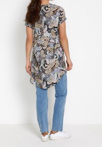 Kvinde iført en kortærmet tunika med paisley-mønster, lys blå jeans og hvide sneakers, stående mod en simpel hvid baggrund.