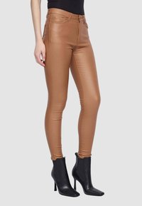 Jeans skinny beige con una texture liscia e lucida e vita alta, abbinati a stivali neri a punta alla caviglia. Progettati per una silhouette aderente.