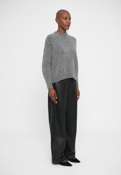 Maglione grigio a maglia con collo tondo e polsini a costine, abbinato a pantaloni neri a taglio largo. Il modello indossa orecchini statement e scarpe nere.