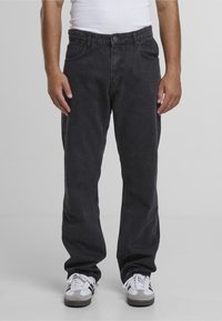 Homme portant un jean droit noir, des baskets blanches avec des bandes noires et un débardeur blanc, debout devant un fond gris uni.