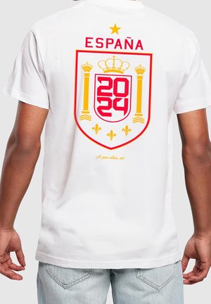Osoba nosząca białą koszulę z czerwono-żółtym emblematem Hiszpanii 2024, koroną, kolumnami i gwiazdą na plecach, powyżej jasnoniebieskich dżinsów.