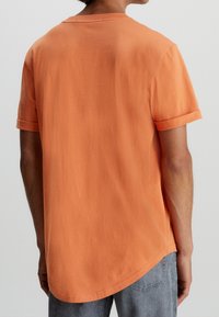 Kortärmad, orange bomulls-T-shirt med rundad nederkant och uppvikta ärmar, visad bakifrån, som visar en slät yta.