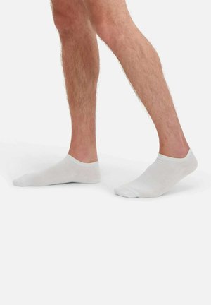 Benen met witte onzichtbare sokken, één voet plat op de grond en de andere licht opgetild op de tenen, tegen een effen witte achtergrond.