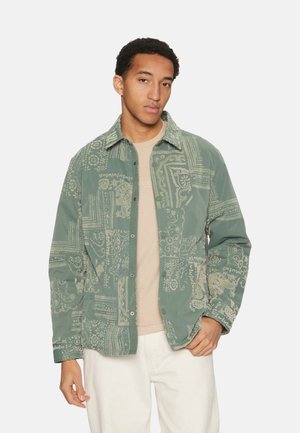 Chaqueta verde con un patrón que presenta un cierre de botones en la parte delantera, un cuello y diseños intrincados en un tono más claro. Llevada sobre una camisa beige.