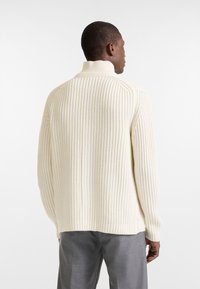 Maglione crema testurizzato con colletto alto e design a costine, dotato di maniche lunghe e vestibilità comoda. Indossato sopra pantaloni grigi.