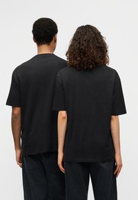 Μαύρα βαμβακε ρά μπλουζάκια, κοντά μανίκια, χαλαρή εφαρμογή, ορατές ραφές, φορεμένα από δύο άτομα από πίσω, χωρίς εμφανή λογότυπα ή σχέδια.