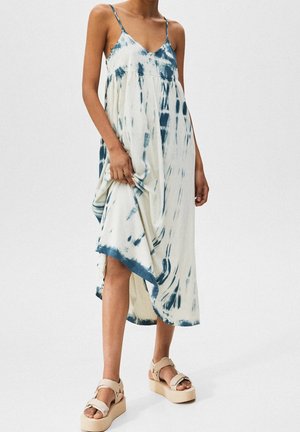 Kobieta w bezrękawną białą i niebieską midi sukienkę tie-dye na cienkich ramiączkach, trzymająca dół sukienki, w beżowych sandałach na platformie na białym tle.