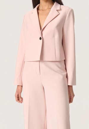 Woman wearing a pale pink cropped blazer with a single button and matching high-waisted wide-leg trousers.