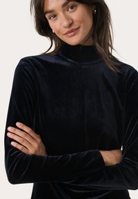Schwarzes Samt-Mock-Turtleneck-Oberteil mit langen Ärmeln, das eine weiche Textur und eine körpernahe Silhouette aufweist. Die Arme sind zum entspannten Aussehen verschränkt.
