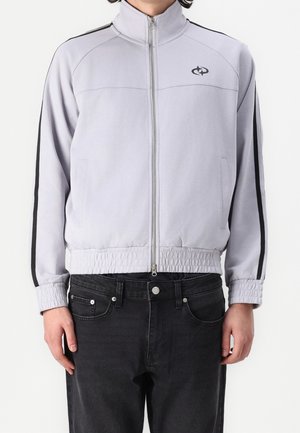Light gray zip-up jacket with high collar, black side stripes, elastic waistband, and side pockets, paired with black jeans.