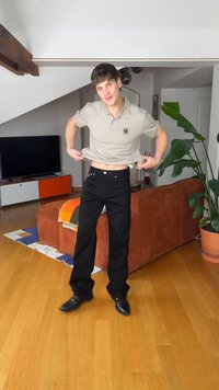 Νεαρός άνδρας σηκώνει μπεζ πόλο πουκάμισο στο σαλόνι με πορτοκαλί καναπέ, ξύλινο πάτωμα, πράσινο φυτεμένο φυτό και τηλεόραση στο φόντο.