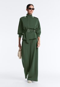 Kvinde iført mørkegrøn jakke med bælte og vidde bukser, der holder en beige taske og har sorte solbriller på, gående på hvid baggrund.