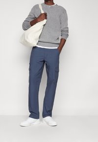 Harmaa teksturoitu villapaita, siniset taskuhousut sivutaskuilla, valkoiset tennarit ja kermaan vivahtava kangaskassi. Yksivärinen tausta.