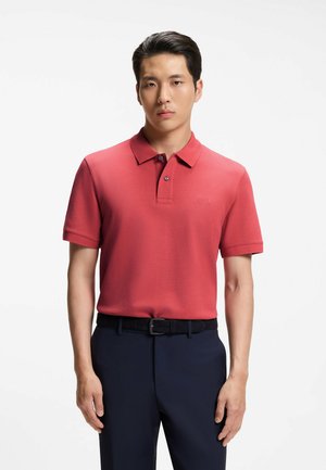 Uomo che indossa una polo rossa a maniche corte e pantaloni blu scuro, in piedi davanti a uno sfondo bianco uniforme.