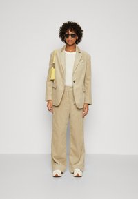 Beige kordfløyelsdress med enkelknapp blazer og bukser med snøring, kombinert med en hvit t-skjorte og en gul håndveske, satt mot en lys bakgrunn.