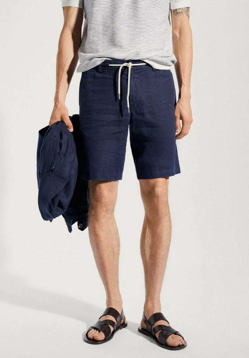 Shorts en lin bleu marine avec une taille à cordon, associés à une chemise rayée. Le modèle tient une veste assortie bleu marine. Des sandales noires complètent le look.