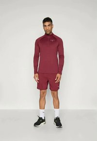Bordeaux treningsoverdel med kvart glidelås og lange ermer, kombinert med bordeaux shorts. Brukt med sorte joggesko og hvite sokker.